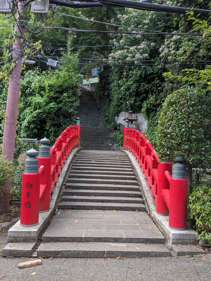 Eine asiatische Brücke mit An- und Abstieg. Rotes Geländer.
Über sie führt eine Treppe in bewaldetes Gebiet dahinter.