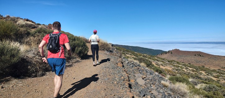 Group of runners on a highland dry rocky path, under a clear blue sky. In the distance, low cloud cover is seen from above.