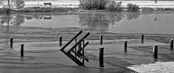 Schwarzweißfotografie im Format 21:9 waagerecht.
Winter an einem Badesee. Es liegt wenig Schnee, ist aber nicht kalt genug für eine geschlossene Eisdecke.
Im Vordergrund ein Badesteg mit Treppe. Das Wasser steht so hoch, dass die Steegfläche gerade unter Wasser ist und die Handläufe der beiden Treppengeländer aus dem Wasser heraus ragen. Das gegenüberliegende Ufer ist als schmaler Streifen zu erkennen. Dort, links oben, führt eine Treppe aus dem Wasser, neben welcher rechts eine Sitzbank aus Holz steht.