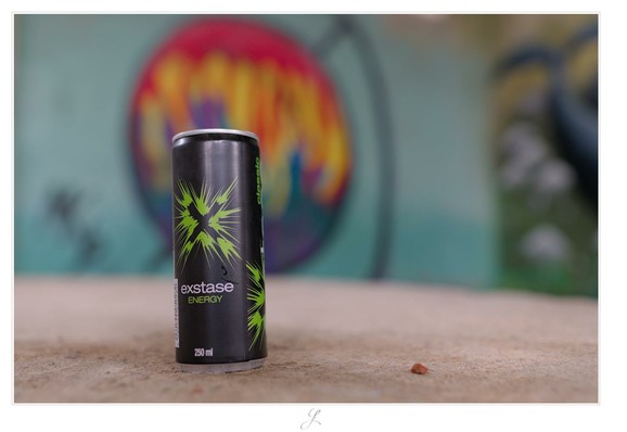 A close-up of a thin tin can. The can is black, an "X" is surrounded by green splotches. Below the label reads "exstase" in silver and "engergy" in green. At the bottom "250ml" can be read. In the blurred background there is a colorful graffiti on a wall. The tin can stands on a concrete surface. Just the can is in focus.

AI disclaimer: Using my work, its meta data, written or derived description to create media with or train AI based systems is prohibited.