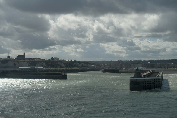 Photograph of the seafront of Dieppe seen from a departing ferry. The surface of the sea is green and rugged, slightly swirly.
The sky, taking nearly half of the picture, has large clouds but also many bright patches of sunlight.
The city itself is divided by the port in a left half with cliffs and a church on top and an urban right half with a beach and the seafront buildings.
A long concrete pier extends towards the viewer from the port.