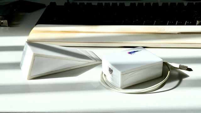 Two hand-made white acrylic enclosures on a white desk. One a right-triangular prism, and one a simple-looking rectangular prism with rounded corners. The former is otherwise featureless, but the latter has a splash of blue and red in the form of a tiny logo on its top side near the top right corner. It also has an ex-iPhone white USB cable loosely coiled around its base, and its side-access RJ45 socket is clearly visible. A soft daylight casts vertical blinds shadows across them, giving them a classic "Gangster film" chic. It's Boxie with the moxie and sharp-angled Sam!