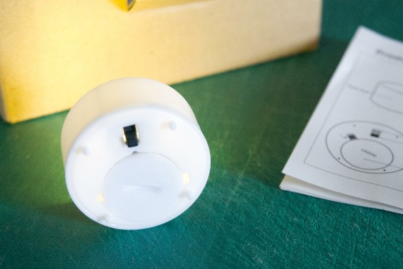 A close-up of the underside of a white LED waxin light on a green background. The switch and battery compartment are visible. In the background is a cardboard box, and to the right is a manual with a diagram.