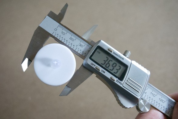 A digital caliper measures the diameter of a white LED waxin light and displays 36.93 mm on the screen. The caliper is held by one hand.