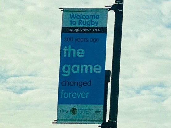 An advertising flag attached to the side of a lamppost. It's blue and white and at the top in smallish writing days "Welcome to Rugby". But, more importantly, in the middle of the flag in large text it says "the game".