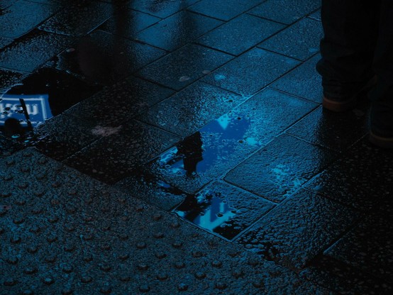 Close-up photo of a wet street reflecting blue color from Time Square's neon signs.
