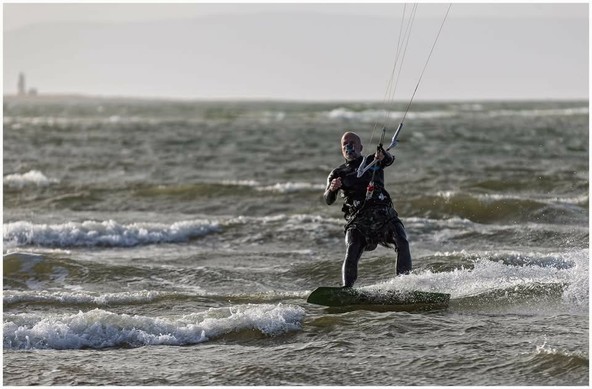 A person kitesurfing on choppy water, wearing a wetsuit and harness, with a kite in the sky above. The background features a distant lighthouse and cloudy sky.