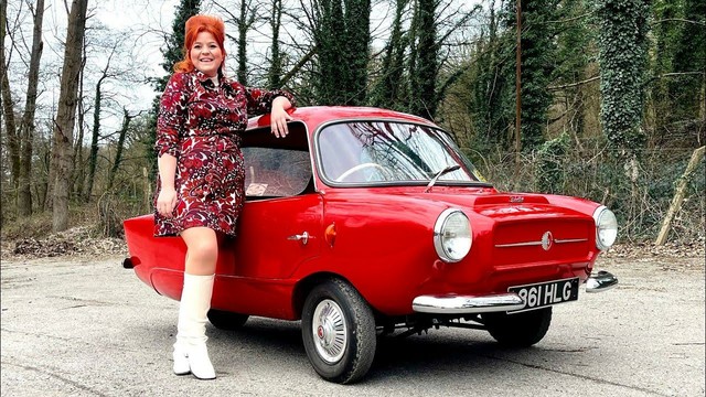 A woman dressed in a 1960s-style red and white patterned dress with white knee-high boots leans on a bright red Peel Trident microcar. The tiny three-wheeled car has round headlights, a minimalist chrome grille, and a UK license plate. The setting is an outdoor wooded area with leafless trees and ivy-covered trunks, giving a retro yet timeless feel.