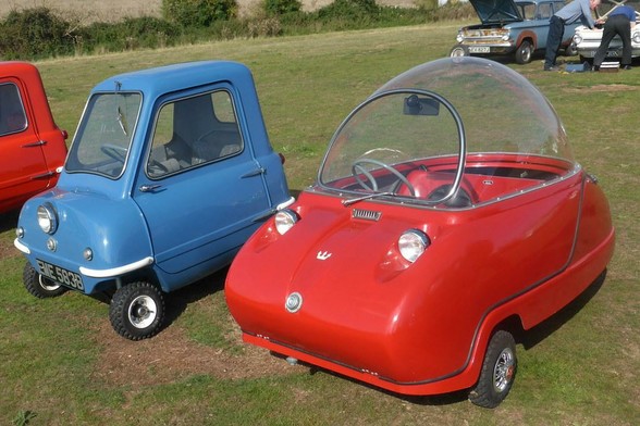 A pair of vintage microcars are displayed on a grassy field. On the left is a blue Peel P50, a tiny three-wheeled car with a single headlight, a compact boxy design, and a UK license plate reading "EME 583B." On the right is a red Peel Trident, featuring a bubble-shaped clear dome canopy, two small headlights, and a futuristic, curvy design. In the background, classic cars with open hoods are being worked on by people, adding to the nostalgic atmosphere.
