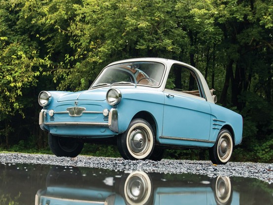 A vintage light blue Autobianchi Bianchina convertible with a white roof is parked on a gravel path near a reflective puddle, surrounded by lush green trees. The car has round headlights, chrome bumpers, and a classic mid-20th-century design. The reflection in the water adds a nostalgic and artistic touch to the scene.
