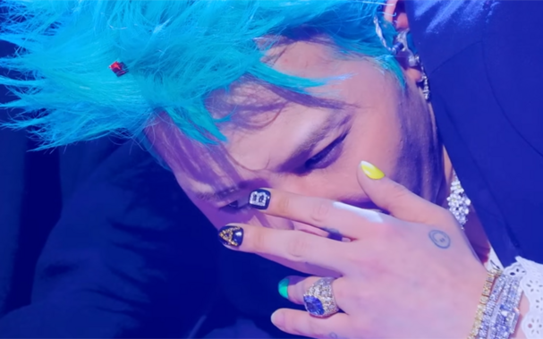 G-Dragon covering his face with one hand and showing his fancy manicure. He has nails done in different colors including yellow, black and teal. Two of his nails have chains or gems glued on to form the the letters A and B. He also has bright blue hair and sparkly jewellery.