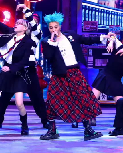 G-Dragon performing on stage while wearing a long red tartan swooshy skirt, black chunky boots, a black cropped jacket with white ruffled cuffs and bright blue hair