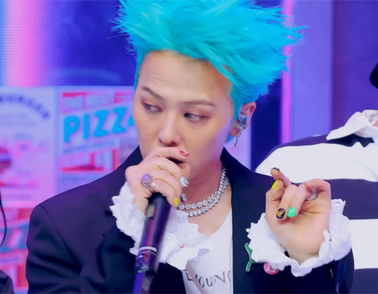 G-Dragon looks to the side while performing on stage and holding a microphone close to his mouth. He is holding up both of his hands as if he wants to show off his fancy manicure, done in lots of bright colors including red, yellow, green. Some of his nails are black or white and have chains or gems glued to them