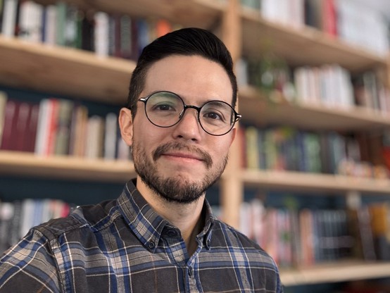 Selfie in front of my bookshelf. I’m wearing a plaid blue shirt and glasses.