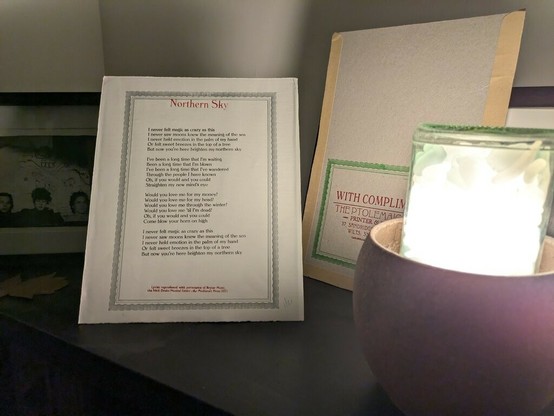 A sheet of printed lyrics are propped on a shelf with a lamp, a card compliments slip, and a framed picture. The sheet has the words Northern Sky printed in red, and the lyrics below in black.