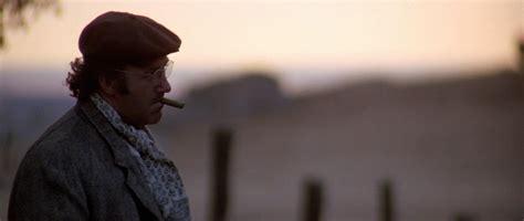 Actor Gene Hackman in the film SCARECROW—shown in a wide shot at dusk wearing round wire-spectacles, a newsboy cap, and smoking a cigar.