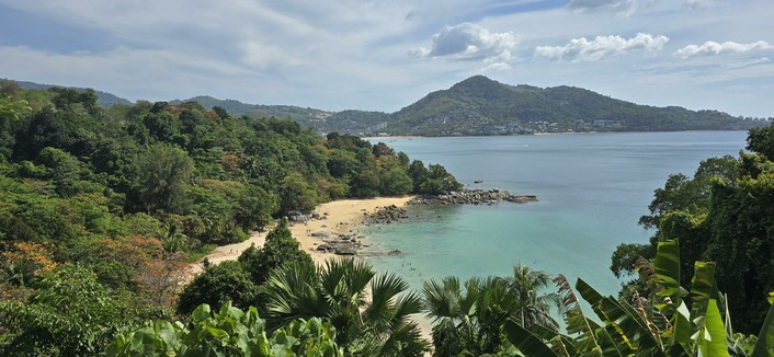 Man sieht eine malerische Küstenlandschaft mit einem üppigen, grünen Wald, der zu einem sandigen Strand führt. Der Strand wird von klarem, türkisfarbenem Wasser gesäumt und es sind mehrere Menschen auf dem Strand und im Wasser zu erkennen.