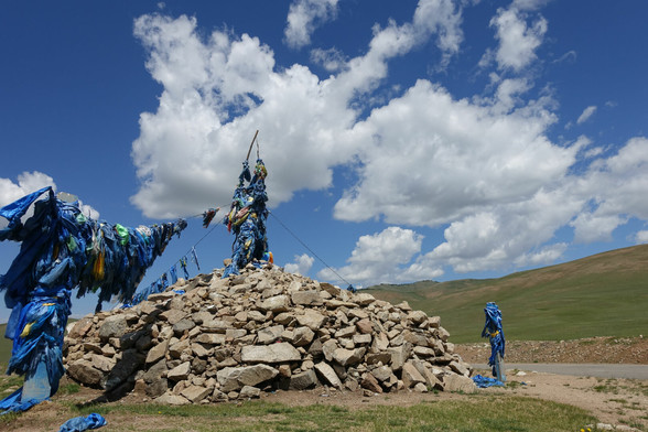 Blauer Himmel mit zerfederten Weissen Wolken, grüne Hügel im Hintergrund, eine Strasse führt von rechts ins Bild. Dominiert wird die Szene von einem großen Steinhaufen aus Bruchsteinen und kleinen Felsen. darin steckt ein Stock der mit blauen Tüchern umwickelt ist, der mit drei Seilen abgespannt ist, die ebenfalls mit haufenweise Blauen Tüchern verziert sind.