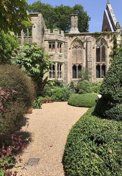 A gravel path leads the way through a collection of topiarized shrubs to the ruin of a stone clad house constructed in the Victorian Gothic tradition. The scene is brightly lit by the midsummer sun. The sky is vibrant blue and cloudless.