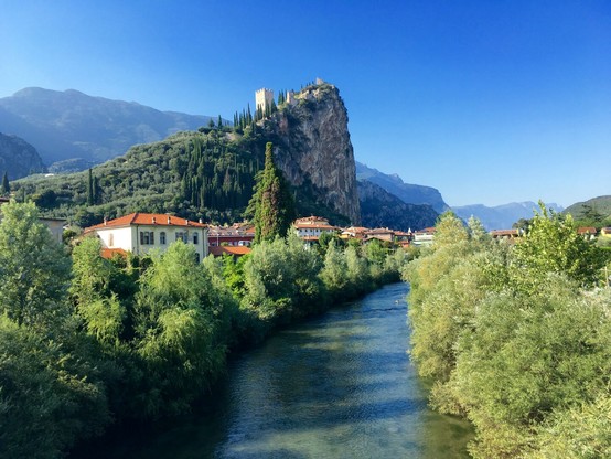Durch die Mitte des Bildes fliest ein begradigter Fluss mit leichter Biegung nach rechts. Am rechten Ufer grün bewachsene Böschung. Am linken Ufer sieht man hinter der Böschung italienische Häuser, überwiegend weiß mit roten Dächern. 

Hinter ihnen ragt ein Berg auf, hinter den Häusern geht er senkrecht hoch, auf der linken Seite steigt er gleichmäßig an und ist mit Bäumen bewachsen. Auf der Kuppe eine Burgrunie, von der vor allem der quadratische Turm aus hellen Steinen noch gut erhalten ist. 

Im Hintergrund eine Bergkette.