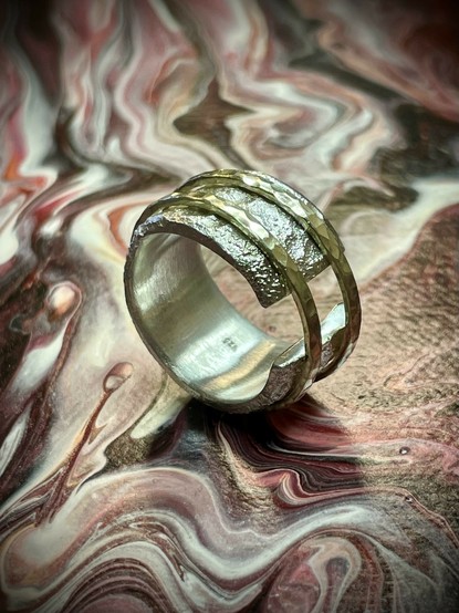 A stylish and edgy ring with three textured bands sits on a colorful, marbled background featuring shades of red, pink, and cream.