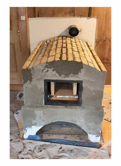 Oven with side walls with abutment bricks and fireclay bricks. Finished fine plastered wall in the background with a round large oven pipe leading into the wall. Old oven door frame refitted in front of the ofen entrance and walled in with rounded lintel brick. Finished arch walled with wedge bricks. Flue gas outlet of the oven on the back left side. Last row with narrow wedge keystones on the arch crown closed. Terracotta tiles in the front covered with brown paper and wooden parquet flooring visible on the let and right of the oven. A old wooden paneling is visible on the left, top and right edges of the photo.