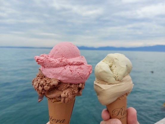 Two ice cream cones are held above a body of water. One cone has a pink scoop and a darker chocolate scoop, while the other features a pale green scoop and a tan scoop. The background shows a cloudy sky and distant mountains.