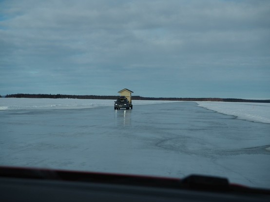 Fotografiert vom Beifahrersitz des Autos. Wir fahren auf der Eisstraße auf der zugefrorenen Ostsee. Uns kommt ein Fahrzeug entgegen, das auf einem Anhänger ein Holzhäuschen transportiert. Das ist eine mobile Sauna, wie sie im Norden von Skandinavien häufiger anzutreffen ist.