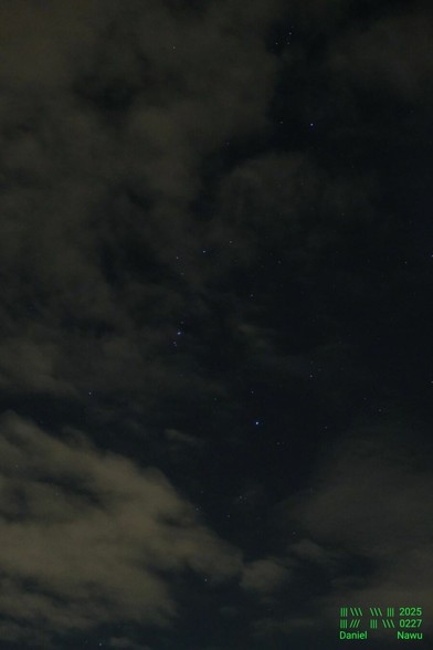 Foto im Hochkantformat. Aufgenommen in der Nacht, daher recht dunkel. Durch ungleichmäßig verteilte Wolken ist das Sternbild Orion auf sehr dunkelblauem Nachthimmel zu sehen. Für mich mutete dieser Moment sehr sinnlich an, wie die Wolken so friedlich dahin zogen und gelegentlich den Blick auf die vielen Sterne freigab.