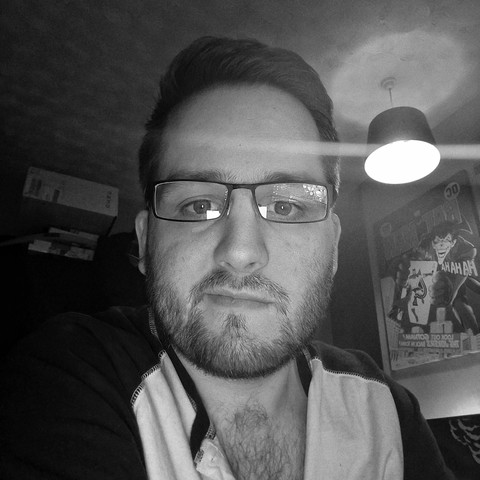 A black and white image of a man sitting at a desk, he has short-ish dark hair and stubble and is wearing glasses, his henley shirt is open slightly and showing some chest hair, behind him are some boxes on top of a wardrobe and an enlarged version of the cover of Batman issue 251