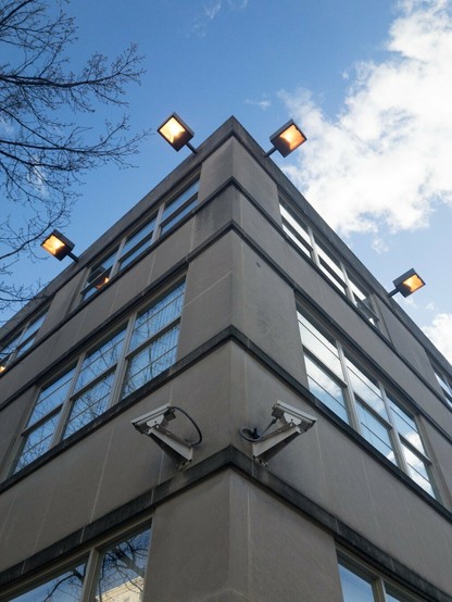 Looking up at the corner of a building with two security cameras and four flood lights turned on during the daytime