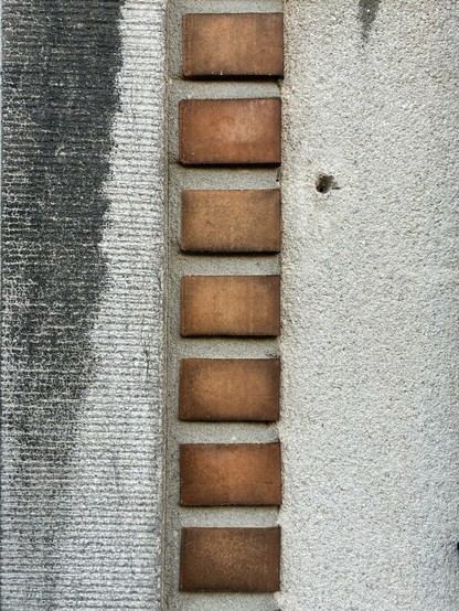 Tussen een stuk blauwsteen en een betonnen muur zit een verticale rij rode bakstenen