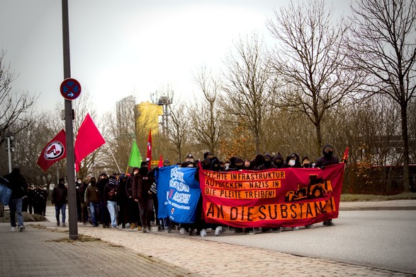Demonstrationszug durch Wellsee. Auf dem Frontbanner steht rechte Infrastruktur aufdecken, Nazis in die Pleite treiben. An die Substanz!. Auf dem Banner ist ein gemalter Radlader an dem eine Antifafahne befestigt ist.