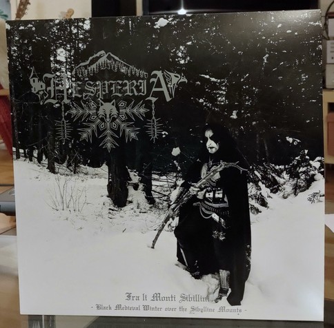 LP cover. Black and white photo of a hooded guy in some kind of armour-like clothing with a black cape, with corpse paint. He stands in the snow in the wood, with trees behind him. His holding a branch or staff, or something like that.

Hesperia logo (with both a big snow crystal, a mountain, a castle, and some creature in it)

"Fra li Monti Sibillini (Black Medieval Winter over the Sibylline Mounts)" on the bottom.