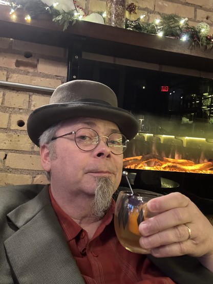 Selfie of a guy with a grey goatee toasting the camera with a cocktail. He is wearing a beaver top hat, glasses, and a grey suitcoat over and orange plaid shirt.