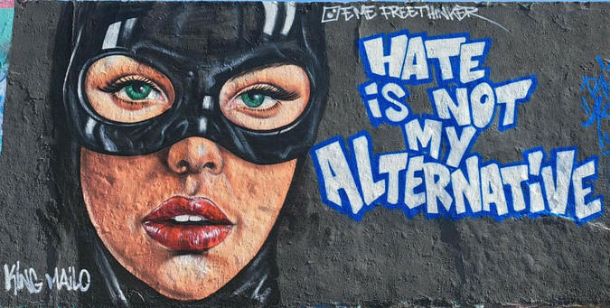 Grafitto. Black background. On the right half in bold white letters 'Hate is not my alternative!' 

Face of a feminine person with green eyes and half open mouth, red lips and a black mask covering the upper part of the face. 

On top a small Instagram logo and  from Eme Freethinker