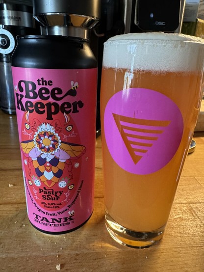 A pink colored can that reads The Bee Keeper pastry sour. The can is standing next to a full glass of cloudy amber beer with a small head