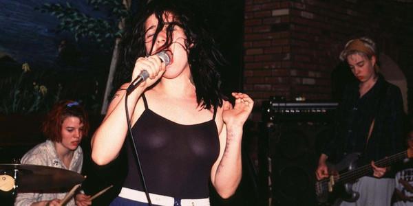 Bikini Kill en concierto a inicios de los 90. De izquierda a derecha, Tobi Vail a la batería, Kathleen Hanna en primer término, con un top negro semi transparente y cantando con el pelo desmelenado y a la derecha, seria y concentrada, Kathi Wilcox, con el pelo corto rubio y tocando el bajo. Discogs.