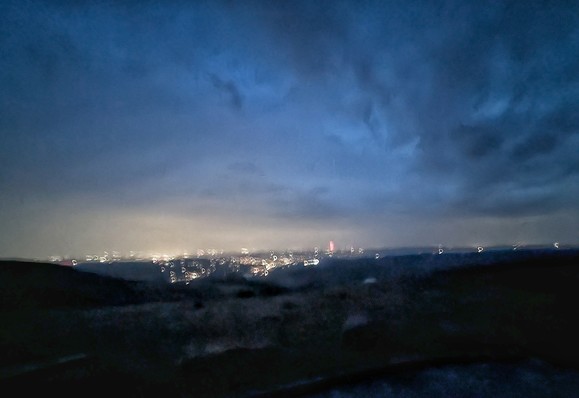 Dark moorland in foreground drops away towards distant night lit city.  Clouds above lit from below.