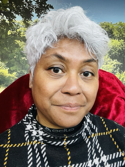 Brown grey-haired woman sitting in a chair with a red blanket wrapped over it. The background is wallpaper with trees and bushes printed on it