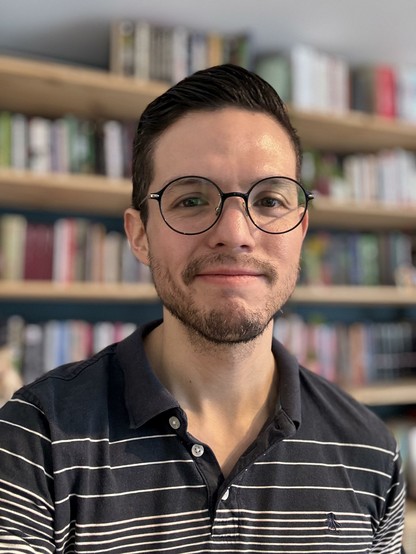 Selfie in front of my bookshelf. I’m wearing a striped blue polo shirt.