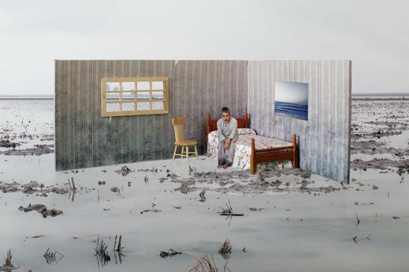 In a room recreated with only two walls, in a theatrical manner, and located in the middle of a swamp, a character sitting on a bed has his feet in the water and looks at the viewer.

---

En una habitación recreada con solo dos paredes, a la manera teatral, y situada en mitad de un pantano, un personaje sentado en la cama tiene los pies en el agua y mira al espectador.