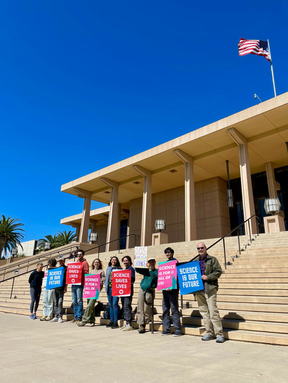 A group of people standing on the steps leading up to a modernist portico, holding signs with pro-science slogans