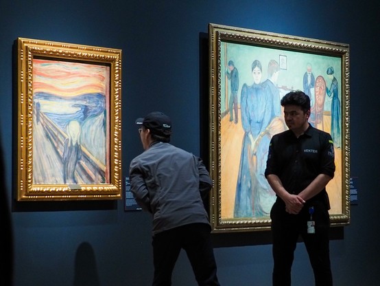 A person wearing a baseball hat looking intensely at Edvard Munch"s famous painting The Scream as close as it was allowed to. Next to him to the right is a security guard watching him like a hawk. Behind the security guard is another, larger painting of Munch's.