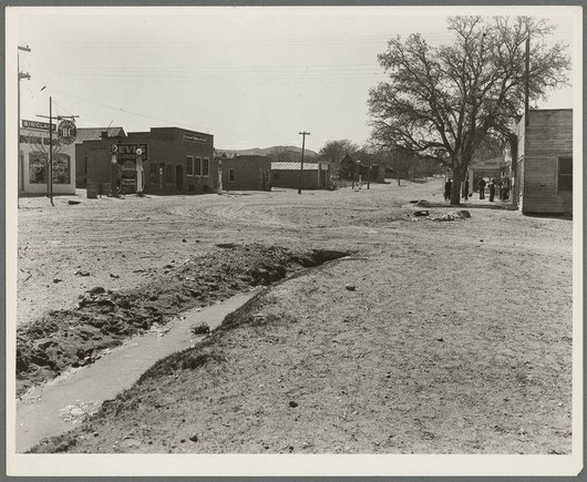 A black and white photograph depicting a dusty rural town street during the early to mid-20th century. The scene includes small, worn-out buildings with various signs such as "EVI" gas station signboard on one of them. A large barren tree stands prominently in front of these establishments, indicating sparse vegetation or an arid climate. In the foreground is a dry irrigation ditch winding through the area, reflecting scarce water resources and possibly agricultural activity once present. Several individuals are visible near the buildings, suggesting minimal pedestrian traffic but human presence within this seemingly quiet locale. The sky above appears clear, with no clouds in sight.
