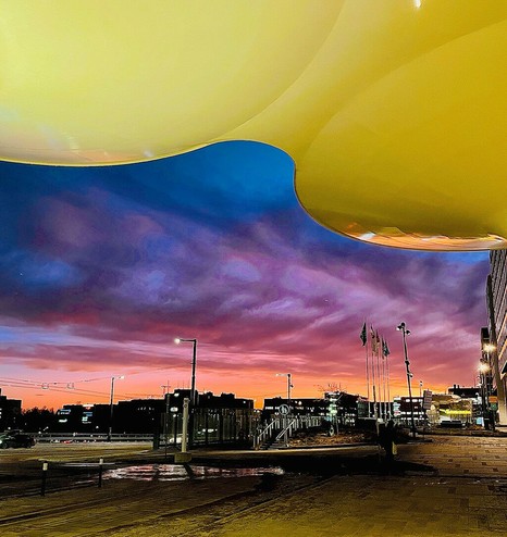A vibrant sunset illuminates the sky in pink and purple hues, framed by a large architectural structure above city street lights below.