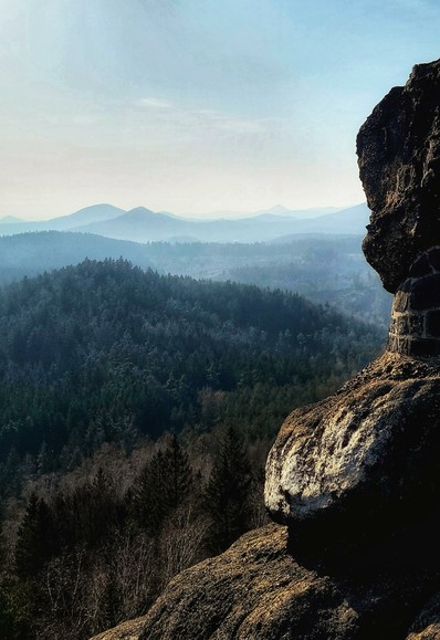 Das Bild zeigt eine weite Landschaft mit Bergen und Wäldern, aufgenommen von einem erhöhten Standpunkt aus. Im Vordergrund befindet sich ein großer, dunkler Felsen, der teilweise im Schatten liegt und dessen Textur deutlich sichtbar ist. Dahinter erstreckt sich ein dicht bewaldetes Gebiet, das in verschiedenen Grüntönen erscheint. Im Hintergrund sind sanfte Hügel und Berge zu sehen, die in einem blassblauen Dunst verschwimmen und sich bis zum Horizont erstrecken. Der Himmel ist hellblau und leicht bewölkt, was der Szene eine friedliche und ruhige Atmosphäre verleiht.