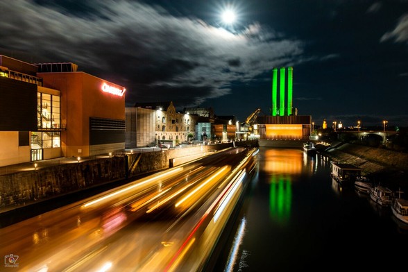 Night scene in a harbor. A boat is approaching the viewer, but only the traces of its yellow lights can be seen (long-exposure effect). Buildings can be seen on the left side above the harbor wall; in front is an illuminated modern, cubic building (Cinemax) and behind it an older brick building. On the right side of the arm of water are five small boats. In the background at the end of the arm of water is an orange-colored, illuminated building on which three tall, slender and round towers - like tall chimneys - can be seen, illuminated in light green. These are also reflected in the dark water. The sky is dark with a few clouds over which the moon shines.