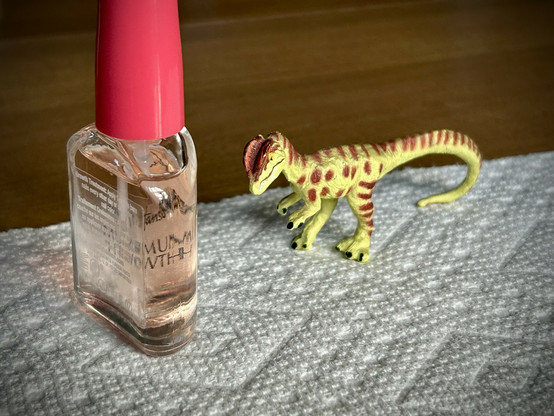 A green Dilophosaurus with brown spots stands beside a bottle of clear nail hardener and awaits their treatment. Both stand on a white paper towel that is itself atop a brown, wooden table.