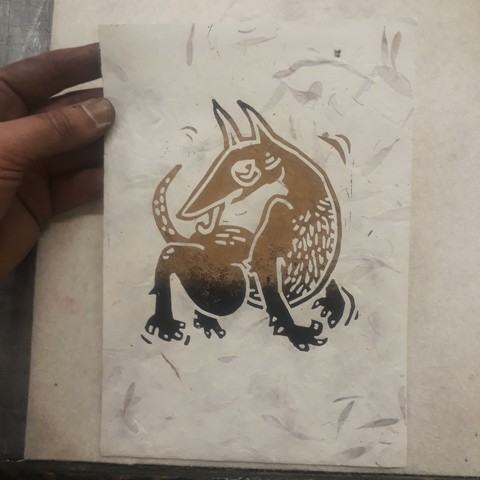 Ma grosse main pleine d'encre tient une feuille de papier incrustée de feuilles d'arbre. Dessus est imprimée une lino de chien bizarre, type naif médiéval, qui se lèche le bas du dos en gigotant. Ses pattes et le haut de ses oreilles sont noires, le reste est doré.